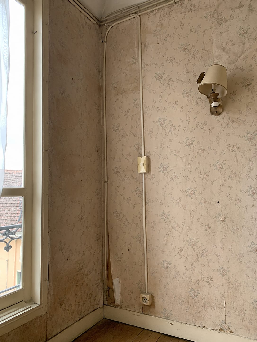 Angle de chambre avant travaux : papier peint défraîchi, câbles en goulotte apparente, interrupteur en saillie jauni et applique cassée.