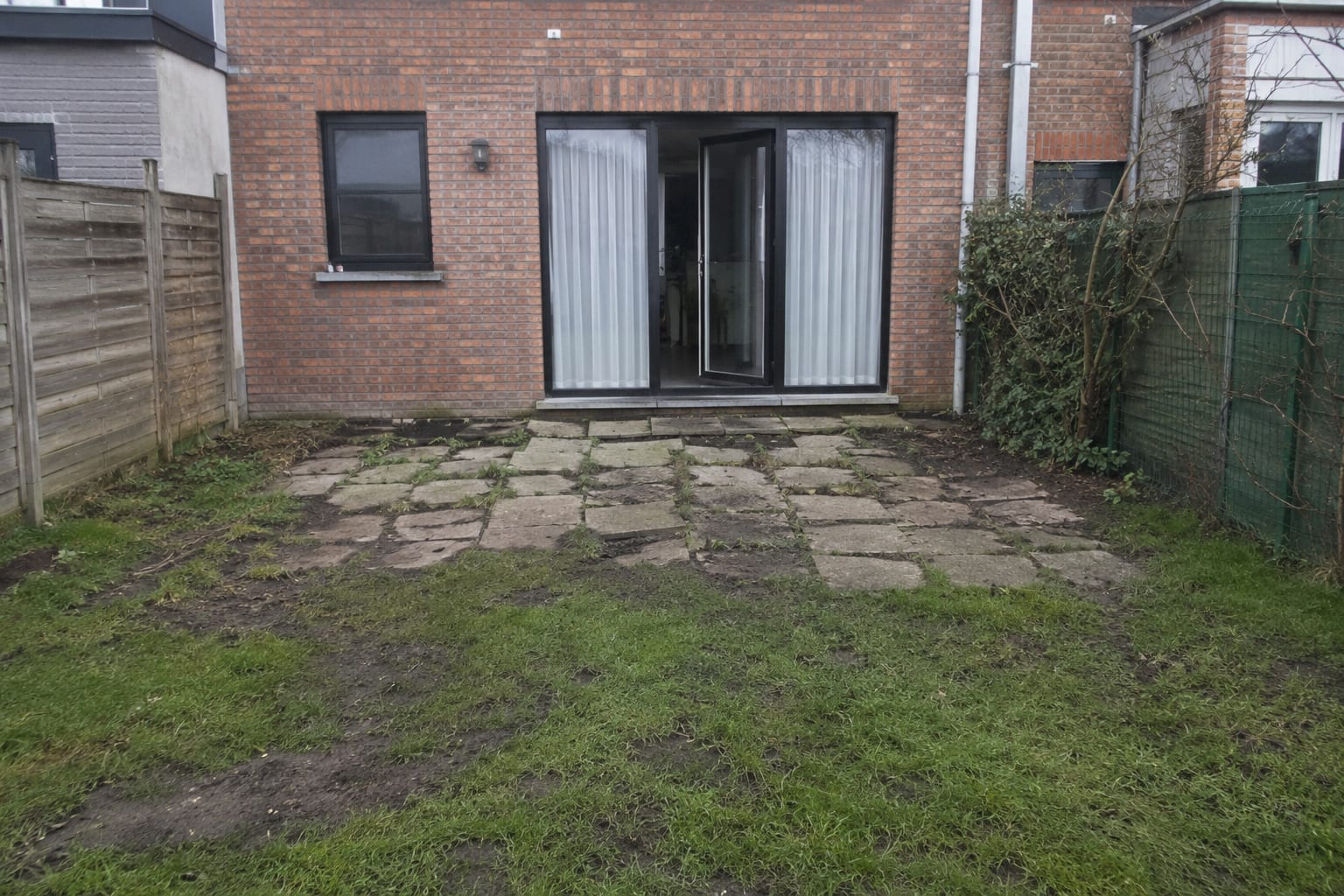 Arrière de maison avant travaux : pelouse abîmée et boueuse, vieilles dalles béton descellées et de guingois devant la porte-fenêtre.