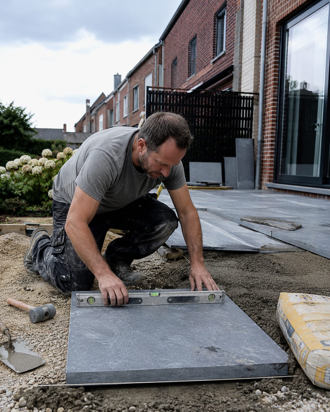 Julien Dethier, maçon, agenouillé sur un chantier de terrasse : il pose une dalle de pierre bleue au niveau à bulle, truelle et maillet à portée de main.